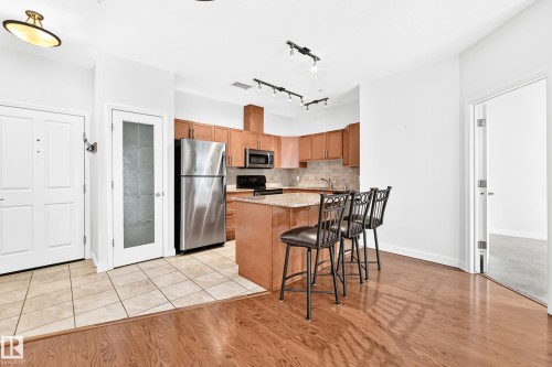 Open-concept kitchen featuring light-toned cabinetry, stainless steel appliances, and a breakfast bar with stone countertops - #307 1003 Gault Bv Nw, Edmonton, AB - Indoor Photo Showing Other Room