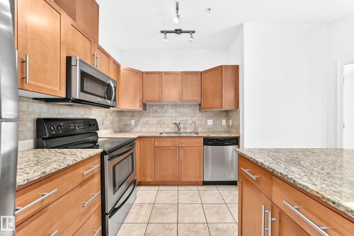 Kitchen featuring wood cabinetry, light-toned countertops, stainless steel appliances, and a tiled backsplash - #307 1003 Gault Bv Nw, Edmonton, AB - Indoor Photo Showing Kitchen