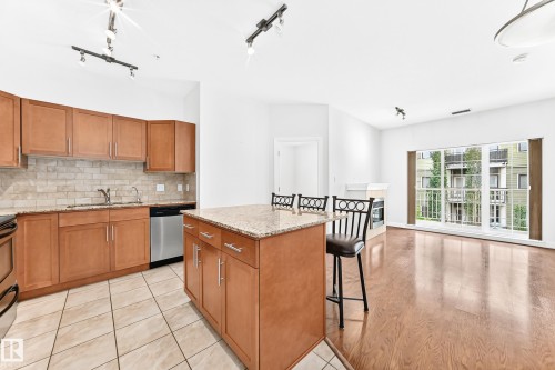 Open-concept living area featuring an island kitchen with wood cabinetry, light stone countertops, and a tiled backsplash - #307 1003 Gault Bv Nw, Edmonton, AB - Indoor Photo Showing Kitchen