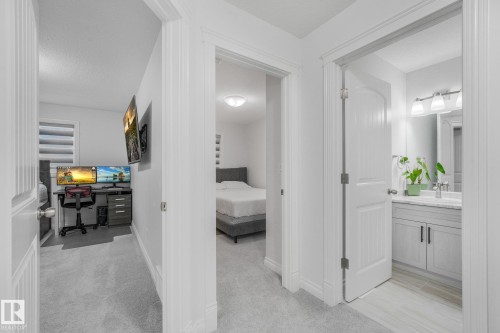 Gray carpeted hallway with white walls and trim, leading to a bathroom with a vanity and a separate room with a bed - 12843 67 Street Nw, Edmonton, AB 