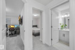 Gray carpeted hallway with white walls and trim, leading to a bathroom with a vanity and a separate room with a bed - 