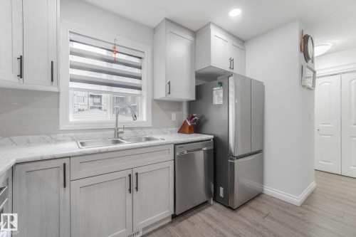 Kitchen featuring white shaker-style cabinetry, stainless steel appliances, a double basin sink with a gooseneck faucet, light-toned countertops, and wood-finish flooring - 12843 67 Street Nw, Edmonton, AB 