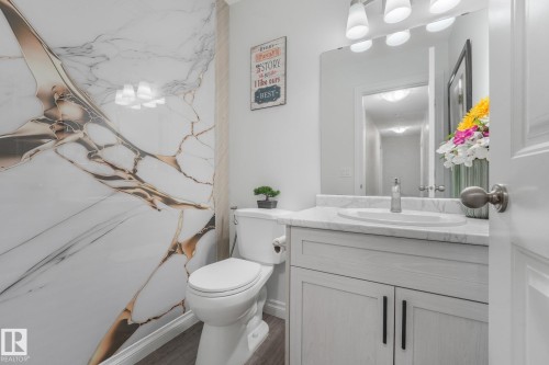 Vanity with a white countertop and integrated sink, under-sink cabinetry with black hardware, wall-mounted mirror, and a wall panel with gold-toned veining - 12843 67 Street Nw, Edmonton, AB 