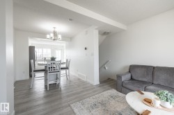 Open-concept living area featuring light wood-finish flooring, white walls, a staircase with white railing, and a dining area with an overhead chandelier - 