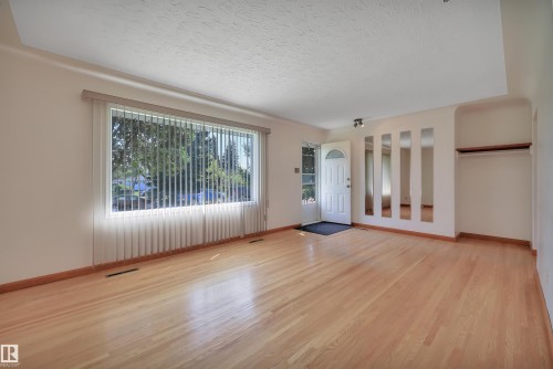 Expansive room featuring light wood-finish flooring, a large window with vertical blinds, a white paneled entry door, and wall-mounted decorative mirrors - 13416 102 Street Nw, Edmonton, AB 