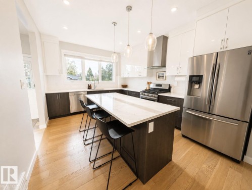 Kitchen featuring a large island with seating, stainless steel appliances, white cabinetry, dark wood-finish lower cabinetry, and light wood-finish flooring - 10516 76 Street Nw, Edmonton, AB 