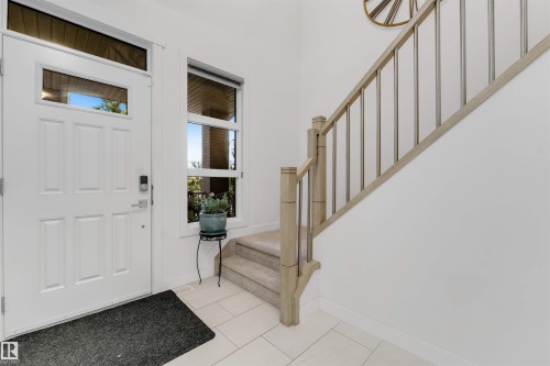 White paneled entry door with transom and sidelight, tile flooring, and carpeted stairs with a wood and metal railing - 718 Charlesworth Way Sw, Edmonton, AB 