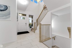 Vaulted ceiling with high windows, white paneled entry door with transom window, light-toned wood-finish staircase with spindles, and rectangular tile flooring - 