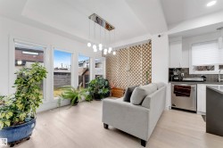 Open-concept living area featuring light wood-finish flooring, multiple large windows, a modern linear chandelier, and integrated kitchen cabinetry with a dark tile backsplash and stainless steel appliances - 