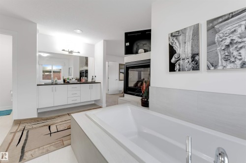 White soaking tub with deck-mounted chrome faucet, light-toned wall tile, and a built-in fireplace - 718 Charlesworth Way Sw, Edmonton, AB 