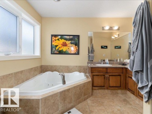 Corner jetted tub with tile surround, adjacent window, and a vanity with a single sink, wood-finish cabinetry, and a large mirror - 496 Ravine Court, Devon, AB 