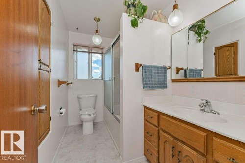 Bathroom featuring a wood-finish vanity with a white countertop and integrated sink, a mirrored medicine cabinet, a toilet, a shower enclosure with a glass door, and a window with blinds - 11415 56 Avenue Nw, Edmonton, AB 