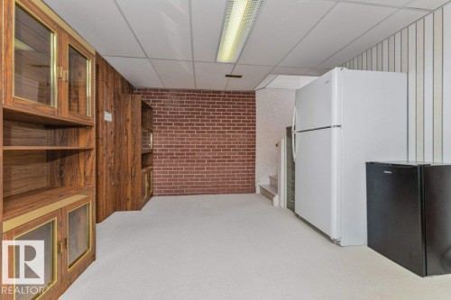 Lower-level room featuring wood-finish cabinetry with glass inserts, exposed brick accent wall, drop ceiling, and carpet flooring - 11415 56 Avenue Nw, Edmonton, AB 