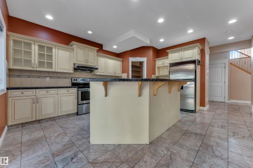 Kitchen featuring an island with a dark countertop, stainless steel appliances, cream cabinetry with glass-front uppers, and tiled flooring - 3455 29 Street Nw, Edmonton, AB 