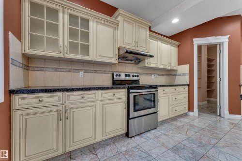 Kitchen featuring cream-toned cabinetry with glass-front uppers, stainless steel range, dark countertops, tiled backsplash, and patterned floor tiles - 3455 29 Street Nw, Edmonton, AB 