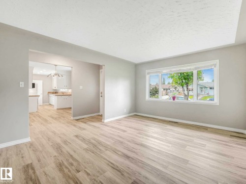 Light wood-finish flooring throughout, bright window, and an open layout with a doorway to the kitchen featuring white cabinetry and light countertops - 15916 88B Avenue Nw, Edmonton, AB 