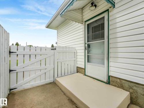 White picket fence with gate, concrete patio, white vinyl siding, exterior lighting fixture, and a storm door - 15916 88B Avenue Nw, Edmonton, AB 