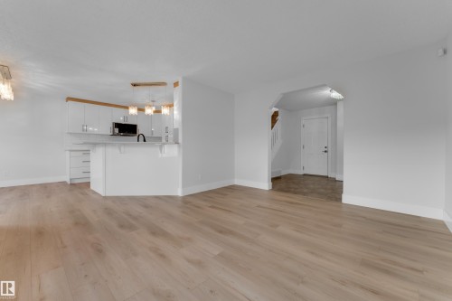 Light wood-finish flooring throughout, white kitchen cabinetry with a light-toned wood accent, a white kitchen island with counter seating, pendant lighting fixtures, and an arched entryway - 3003 25 Avenue Nw, Edmonton, AB 
