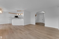 Light wood-finish flooring throughout, white kitchen cabinetry with a light-toned wood accent, a white kitchen island with counter seating, pendant lighting fixtures, and an arched entryway - 