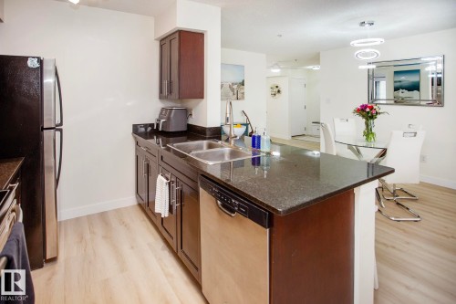 Kitchen featuring dark wood cabinetry, stainless steel appliances, a double basin sink, and wood-finish flooring - #201 1070 Mcconachie Bv Nw, Edmonton, AB 