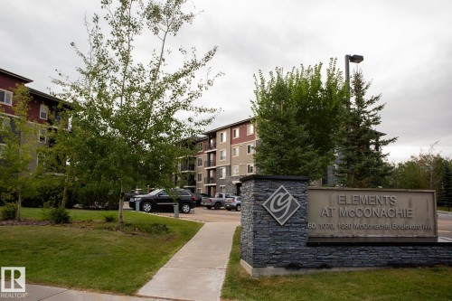 Building exterior featuring a stone-clad monument sign, mature trees, and a paved walkway - #201 1070 Mcconachie Bv Nw, Edmonton, AB 
