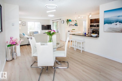 Wood-finish flooring throughout, modern circular pendant lighting, built-in kitchen cabinetry with stainless steel appliances, and a kitchen island with a black countertop - #201 1070 Mcconachie Bv Nw, Edmonton, AB 