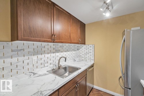 Wood-finish cabinetry, stainless steel sink with a high-arc faucet, white marble-look countertops, and a white tile backsplash with contrasting grout - #1307 10045 117 St Nw, Edmonton, AB 