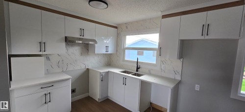 Modern kitchen featuring white cabinetry with dark hardware, a stainless steel range hood, stone-finish backsplash, and wood-finish flooring - 13812 63 Street Nw, Edmonton, AB 