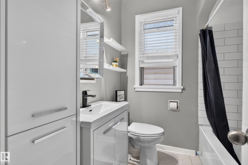 Modern white vanity with integrated sink and matte black faucet, wall-mounted mirror, white subway tile shower surround, and white window blinds - 11406 83 Street Nw, Edmonton, AB 
