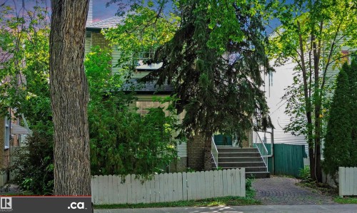 White picket fence with a paver driveway, mature trees, and an exterior staircase with metal railings - 10941 81 Avenue Nw, Edmonton, AB 