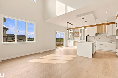 Open-concept living space with light wood-finish flooring, high ceilings, large windows, and a modern kitchen featuring an island, white cabinetry, and a patterned backsplash - 10458 104 Street, Morinville, AB 
