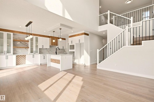 Open-concept living space with light wood-finish flooring, a contemporary kitchen featuring an island, and a dual-level staircase with black iron spindles - 10458 104 Street, Morinville, AB 
