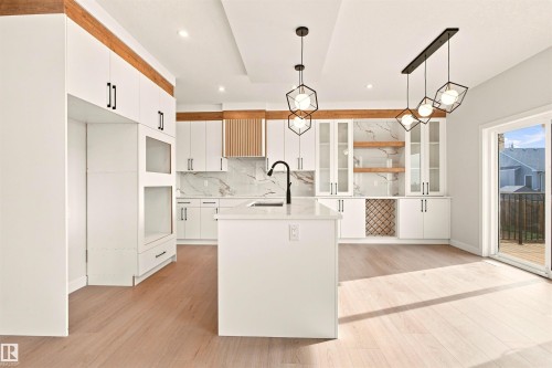 White cabinetry with black hardware, wood-finish flooring, central island with sink, marble-patterned backsplash, and a sliding glass door leading to an outdoor deck - 10462 104 Street, Morinville, AB 