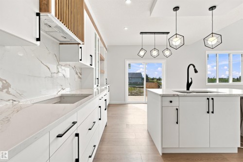 Modern kitchen featuring white cabinetry with matte black hardware, a large island with sink and faucet, marble-look countertops and backsplash, and light wood-finish flooring - 10462 104 Street, Morinville, AB 