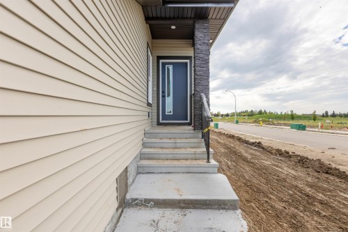 Cream-colored horizontal siding, dark grey stacked stone accent, contemporary dark grey entry door with glass inset, concrete steps, and a dark metal handrail - 10462 104 Street, Morinville, AB 
