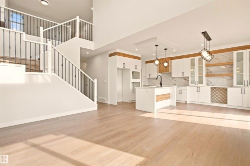 Open-concept living area featuring light wood-finish flooring, a contemporary kitchen with white cabinetry, a kitchen island with sink, black pendant lighting, and a staircase with black metal balusters - 10462 104 Street, Morinville, AB 