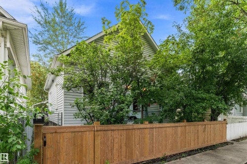White siding exterior with a pitched roof, surrounded by mature trees, featuring a wood privacy fence and a white picket fence - 10816 92 Street Nw, Edmonton, AB 