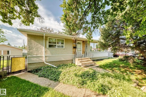 Mid-century modern exterior featuring a stucco finish, a concrete walkway, mature trees, a metal front door with a storm door, and a low-pitched roofline - 15703 84 Avenue Nw, Edmonton, AB - Outdoor