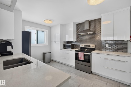 Kitchen featuring white cabinetry, light-toned countertops, a stainless steel range hood, dark brick-patterned tile backsplash, and light gray tile flooring - 13512 111 Street Nw, Edmonton, AB 