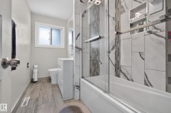 Wood-finish tile flooring, white vanity with integrated sink, white bathtub with a glass shower enclosure, and tiled shower walls with recessed niches - 