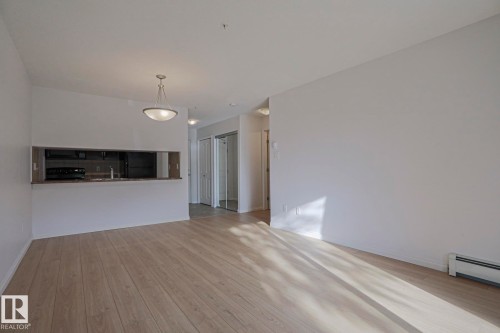 Open-concept living area featuring wood-finish flooring, a contemporary pendant light fixture, and a pass-through window to the kitchen with dark countertops - #108 1204 156 St Nw, Edmonton, AB 