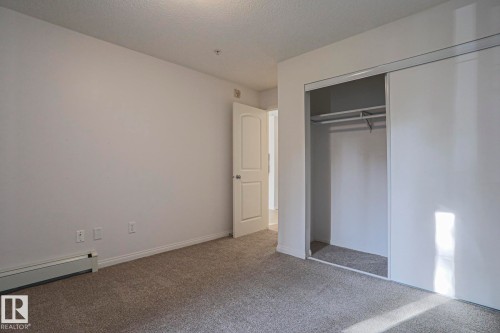 Carpeted room featuring a double-door closet with shelving and hanging rod, white walls, and a baseboard heater - #108 1204 156 St Nw, Edmonton, AB 
