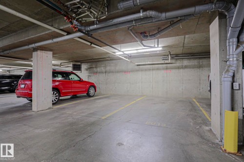 Concrete flooring in a subterranean parking area, featuring exposed overhead piping and fluorescent lighting fixtures - #108 1204 156 St Nw, Edmonton, AB 