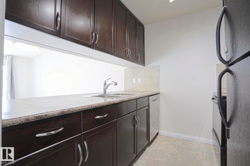 Kitchen featuring dark wood-finish cabinetry, light-colored countertops, dual basin stainless steel sink, and tile backsplash - #108 1204 156 St Nw, Edmonton, AB 