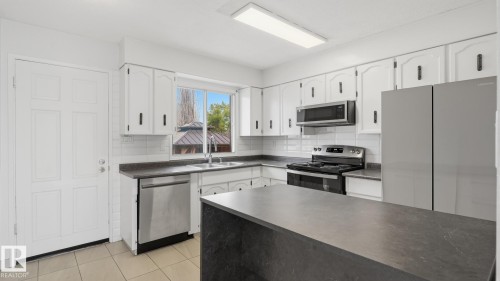 White cabinetry with black hardware, stainless steel appliances, dark countertops, a kitchen island, and tiled flooring - 12208 146 Avenue Nw, Edmonton, AB 