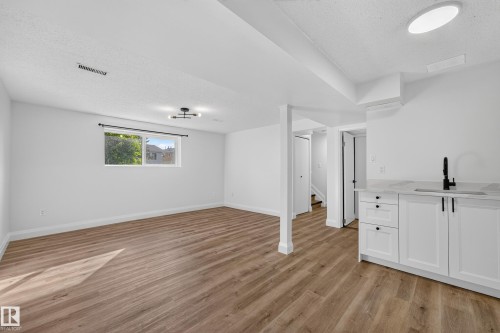 Spacious lower-level room featuring wood-finish flooring, a window, and a wet bar with white cabinetry, a countertop, and a black faucet - 2412 80 Street Nw, Edmonton, AB 