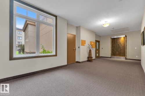 Spacious hallway featuring textured carpet flooring, a large multi-pane window, and a built-in wall water feature - #105 160 Magrath Rd Nw, Edmonton, AB 