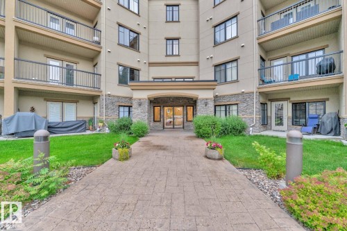 Building exterior featuring stacked stone accents, multiple balconies with black railings, a paved walkway, and a landscaped entryway with green lawn and shrubs - #105 160 Magrath Rd Nw, Edmonton, AB 