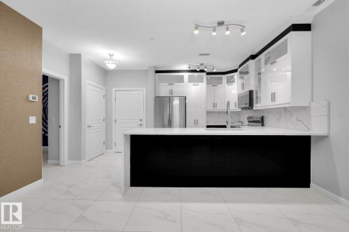 Modern kitchen featuring white cabinetry with black accents, stainless steel appliances, a white countertop with a waterfall edge, and a marble-look tile floor - #105 160 Magrath Rd Nw, Edmonton, AB 