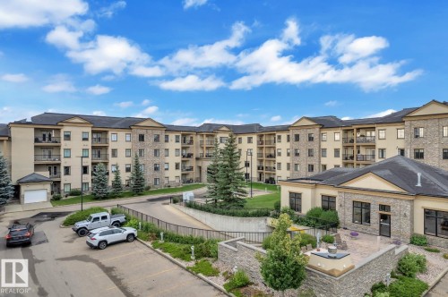 Exterior featuring stone and stucco finishes, multiple balconies, a paved parking area, a manicured lawn, and a stone patio - #105 160 Magrath Rd Nw, Edmonton, AB 